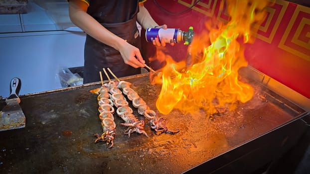 cook grills skewered squid over an open flame using a blowtorch, capturing the lively atmosphere of a bustling street food Thai market with bright flames and freshly prepared seafood.
