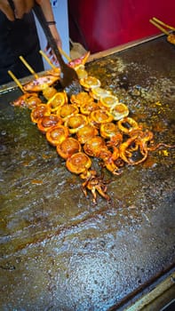cook grills skewered squid over an open flame using a blowtorch, capturing the lively atmosphere of a bustling street food Thai market with bright flames and freshly prepared seafood.