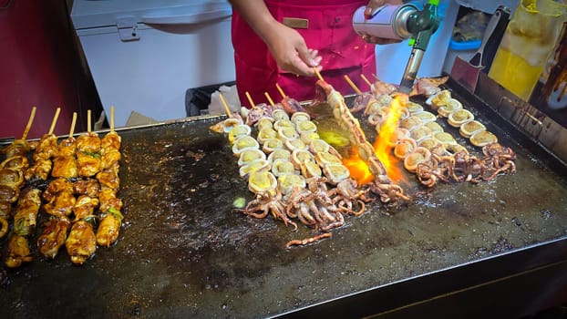 cook grills skewered squid over an open flame using a blowtorch, capturing the lively atmosphere of a bustling street food Thai market with bright flames and freshly prepared seafood.