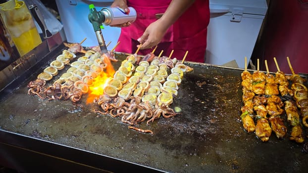 cook grills skewered squid over an open flame using a blowtorch, capturing the lively atmosphere of a bustling street food Thai market with bright flames and freshly prepared seafood.