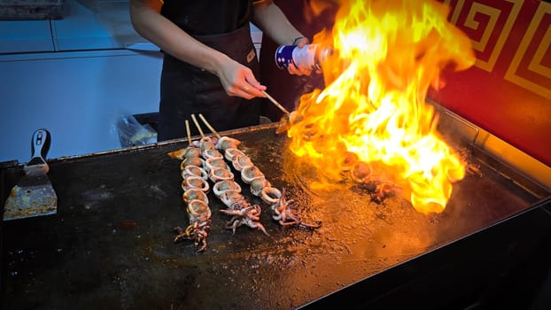 cook grills skewered squid over an open flame using a blowtorch, capturing the lively atmosphere of a bustling street food Thai market with bright flames and freshly prepared seafood.