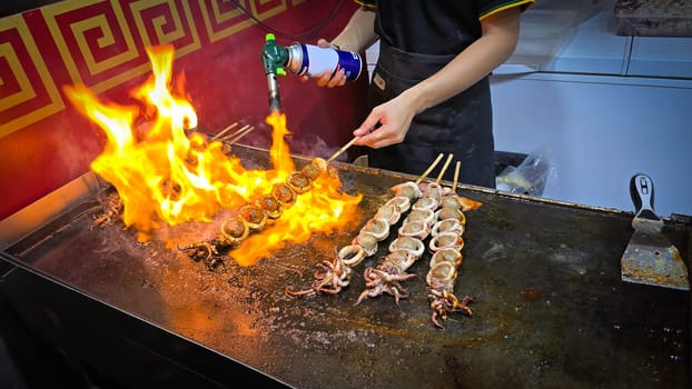 cook grills skewered squid over an open flame using a blowtorch, capturing the lively atmosphere of a bustling street food Thai market with bright flames and freshly prepared seafood.