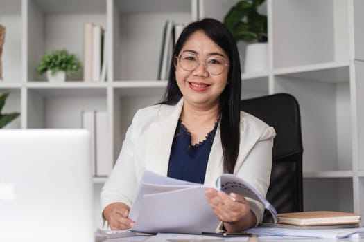 A middle-aged female accountant is sitting at a desk, working on financial documents in a modern office environment.