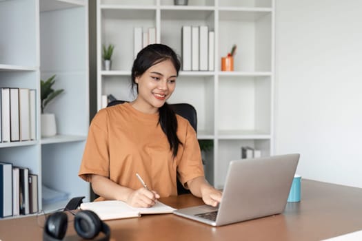 A young girl studies online from home, using a laptop and notebook, in a modern home office environment.