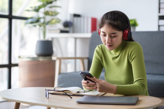Asian woman using smartphone while working with headphones in modern office, concept of multitasking.