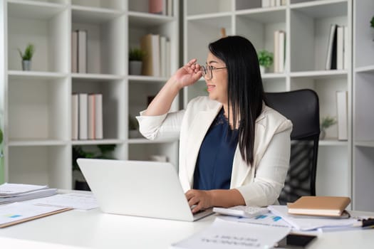 A middle-aged business woman in a modern office, working on a laptop, surrounded by documents and bookshelves.