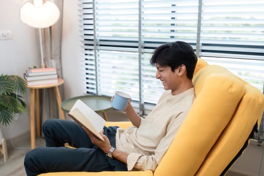 A young man sits comfortably in a modern living room, reading a book and holding a hot beverage, creating a cozy and relaxed atmosphere.