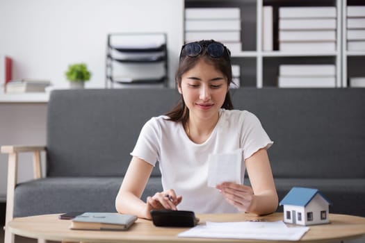A young woman planning her finances for buying a house, calculating expenses with documents and a calculator, sitting in a modern living room.