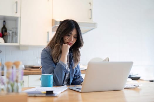 A young freelance woman feeling stressed while working from home, sitting in a modern kitchen with a laptop and coffee mug.