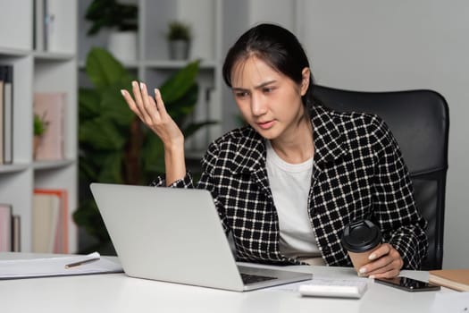 A young accountant feeling stressed while working on a laptop in a modern office setting, holding a coffee cup.
