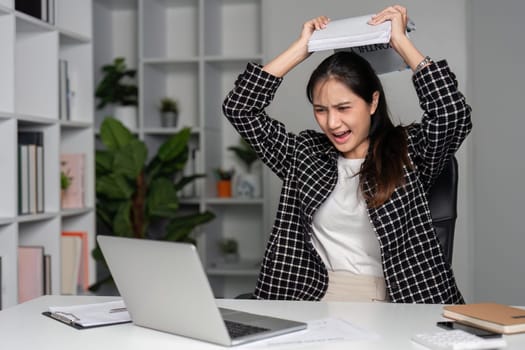 Angry female office worker raising documents in frustration while sitting at her desk with a laptop in a modern office environment.