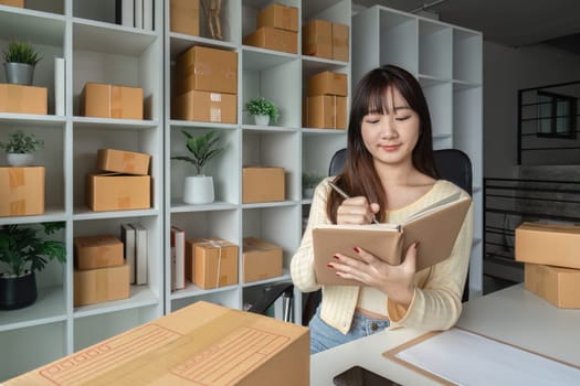 Cute woman taking online sales orders in a modern home office, surrounded by shipping boxes and shelves, writing in a notebook.