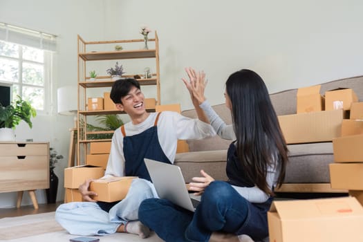 A joyful couple celebrates their successful online sales with a high five, sitting in a modern living room surrounded by shipping boxes and a laptop.