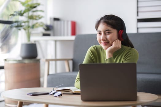 A young woman studies online from home, using a laptop and headphones, seated in a modern living room with a cozy atmosphere.