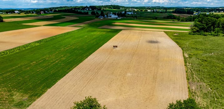 A vast agricultural landscape showcases alternating patches of green and brown fields under a blue sky. A small group of farmers work diligently in the distance, tending to crops.