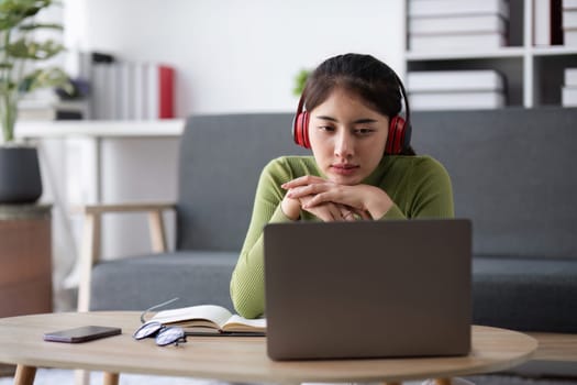 Young woman experiencing stress while working online from home, wearing headphones and using a laptop in a modern living room setting.
