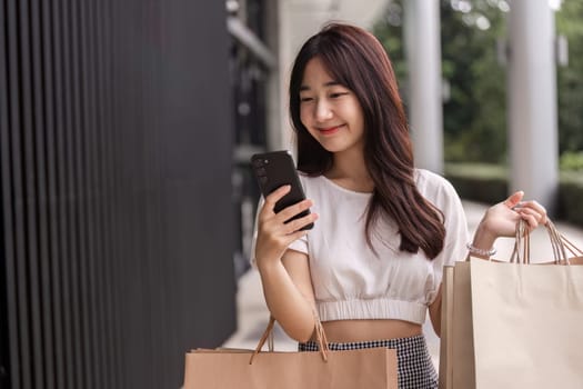 A young woman smiles while shopping, holding a paper bag and using her smartphone in a modern urban environment.