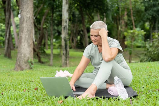 Elderly woman sitting on grass in a park, looking stressed while using a laptop. She is dressed in casual clothing, surrounded by nature.