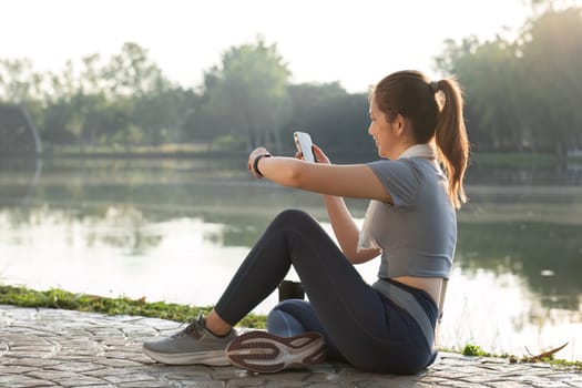 A young woman sits and rests after exercising by a serene lake, checking her smartphone and enjoying the peaceful outdoor environment.