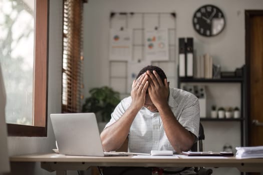 A male office worker experiencing stress and pressure at his desk, surrounded by work materials in a modern office setting.