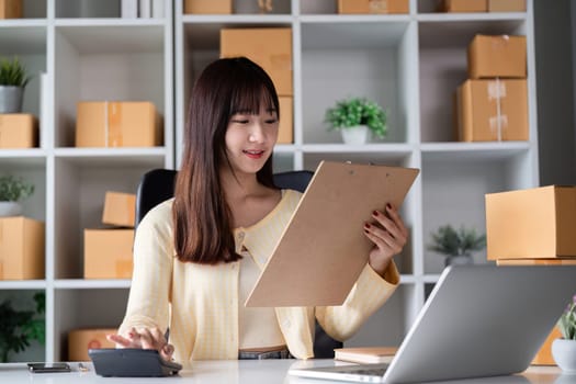 A young female entrepreneur working in a modern home office, managing online sales with a laptop, clipboard, and boxes around her.