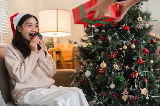 Excited young woman receives a surprise gift from her partner on Christmas Day, sitting by a beautifully decorated tree.
