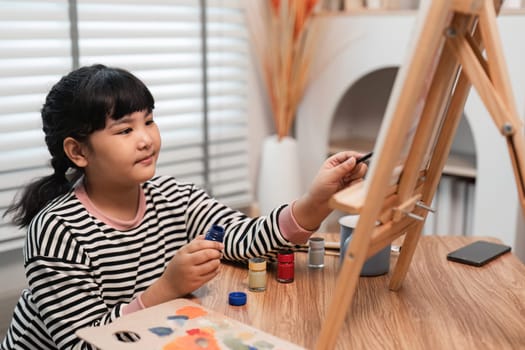 A young girl paints with acrylics on a canvas in a cozy home studio. She is focused and enjoying her creative activity.