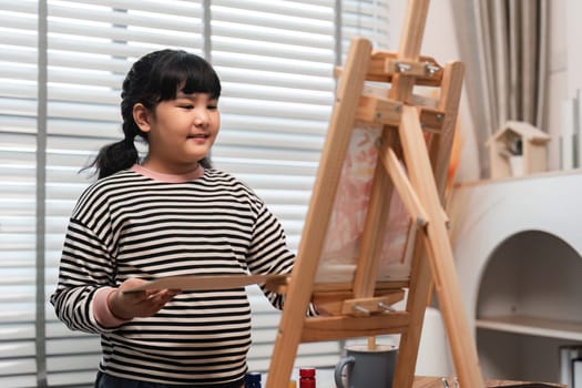 A young girl happily paints with acrylics on a canvas in a well-lit, modern art studio, showcasing her creativity and artistic skills.