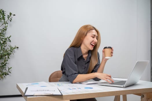 A young female accountant is engaged in financial accounting tasks, using a laptop and enjoying a coffee in a modern office environment.