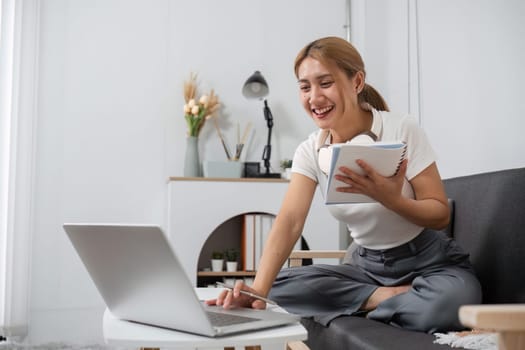 A young woman uses her laptop to watch online entertainment media while sitting on a couch at home, smiling and taking notes.
