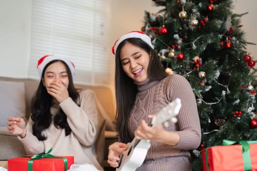 A young female couple enjoys Christmas together, exchanging gifts and playing music by a beautifully decorated tree in a cozy home.