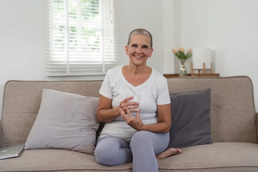 Elderly woman sitting on a sofa, holding a glass of water, smiling, and looking healthy and content in a bright, cozy living room.