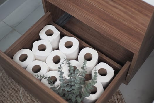 In a wooden drawer, neatly arranged rolls of toilet paper sit beside vibrant greenery
