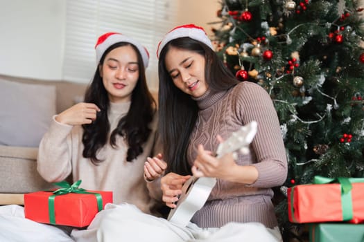 Young female couple enjoying Christmas together, exchanging gifts and playing music by a decorated tree.