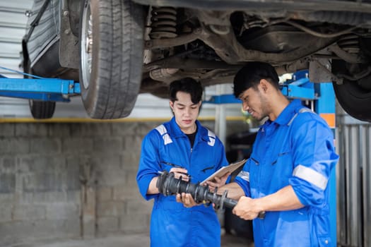 Young man in blue uniform repairing car at auto repair shop, inspecting parts with a mechanic, discussing maintenance and repairs.