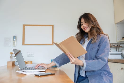 A woman working from home in a dining room, using a laptop and reading a book. Casual and modern workspace setup for remote work.
