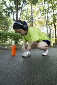 A petite woman prepares for a jog in the park, wearing headphones and athletic gear, with a water bottle nearby.