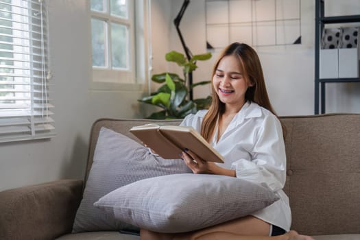 A young woman sits on a couch in the morning, reading a book and relaxing in a cozy home environment.