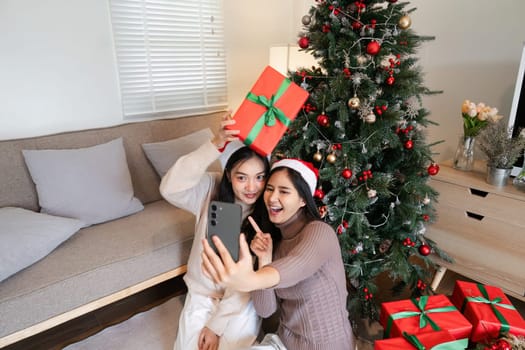 Young female couple joyfully taking selfies at a Christmas party, surrounded by festive decorations and gifts.