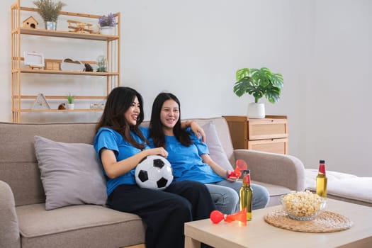 Lesbian couple cheering for football together on a cozy sofa, enjoying snacks and drinks, celebrating sports enthusiasm in a modern home setting.