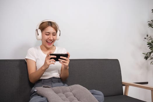 A young woman sits comfortably in a modern living room, wearing headphones and using a smartphone, enjoying a moment of relaxation.