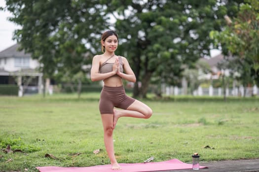 A young woman performs yoga in a park, standing in tree pose on a pink mat, surrounded by lush greenery and trees.