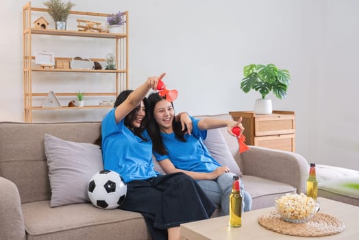 Lesbian couple cheering for football together on a sofa, enjoying game day with snacks and drinks in a cozy modern living room.