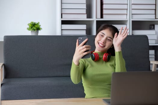 A cheerful young woman in a green sweater relaxes in a modern living room, using her smartphone and wearing red headphones.