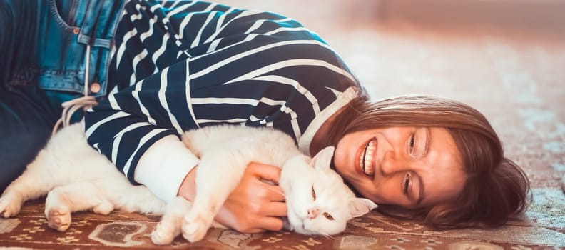 A young cute girl lies on the carpet of the room in an embrace with her white fluffy pet, a woman in a striped sweater smiles and hugs her purring best friend cat, the owner takes care of her kitty.