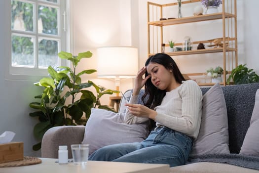 A young woman sitting on a couch, taking medicine while feeling unwell, in a cozy and modern living room with plants and soft lighting.