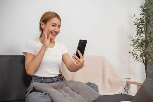 Young woman sitting on a sofa in a modern living room, smiling and waving at her smartphone, enjoying a casual and comfortable moment at home.