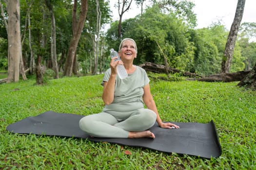 Elderly woman practicing yoga in a park, smiling and holding a water bottle, promoting a healthy and active lifestyle.