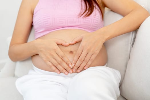 Close up pregnant woman sit on sofa and put hands on her belly with heart shape.