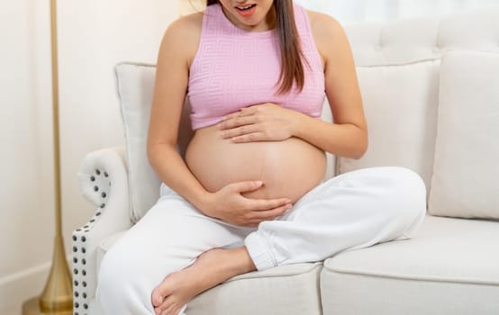 Pregnant woman sit on sofa of her house and look like she feel pain that she touch her belly.